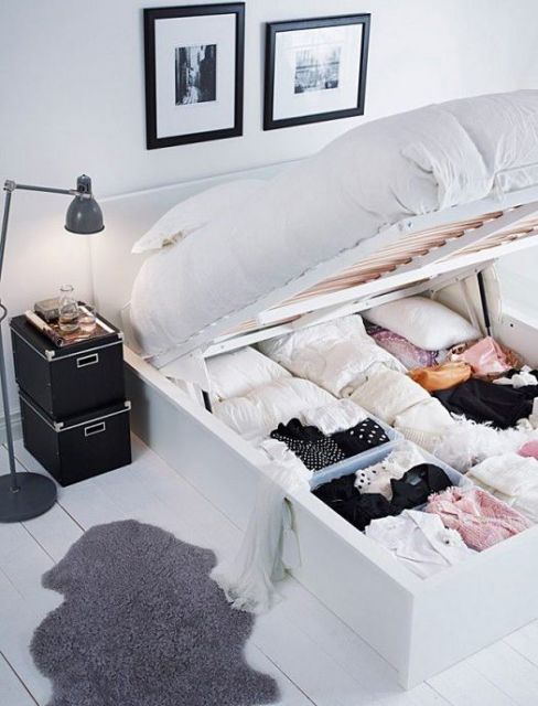 Modelo de cama-baú de MDF branco mostrando roupas pessoais e de cama armazenadas. Ao lado, um criado-mudo de caixas pretas e luminária de chão.