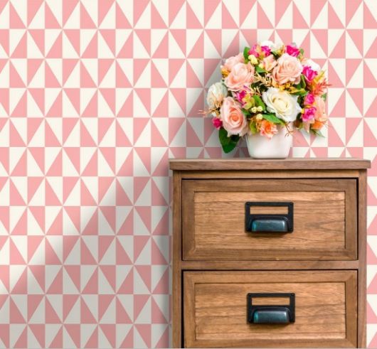Móvel de madeira com vaso de flor em cima e papel de parede com triângulos brancos e rosas.