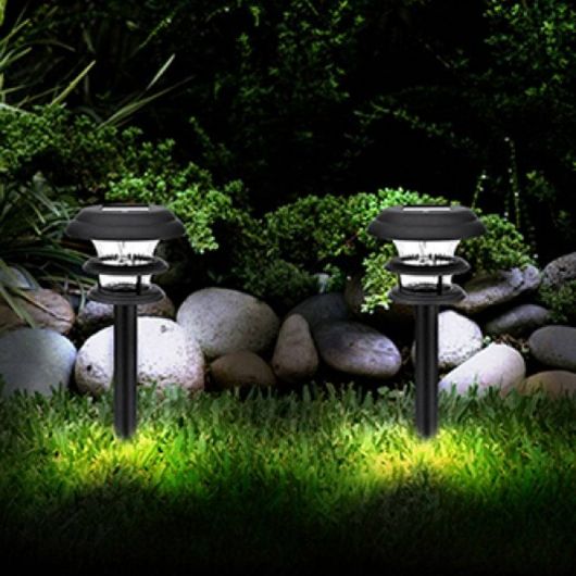 Duas luminárias solares lado a lado em um jardim com grama, árvores e pedras de decoração. 