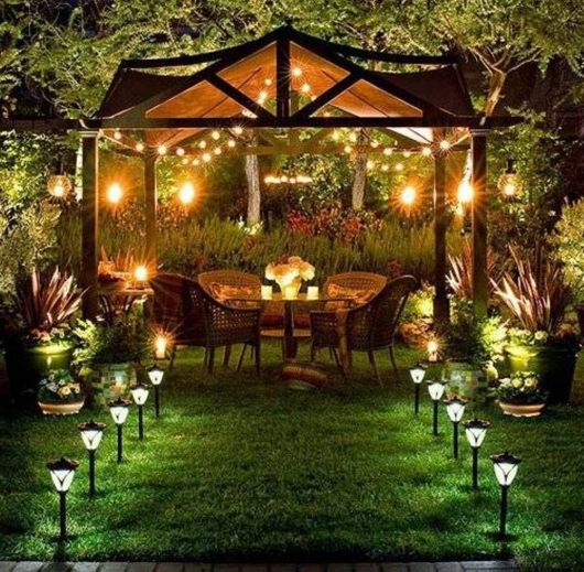 Espaço muito iluminado de um jardim com uma mesa e quatro cadeira. Existem diversas luminárias solares que iluminam o caminho até esse espaço. 