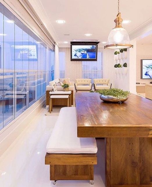 Foto de varanda gourmet espaçosa e clara com uma mesa da jantar grande e sofás ao fundo próximos a um televisor instalado no teto. 