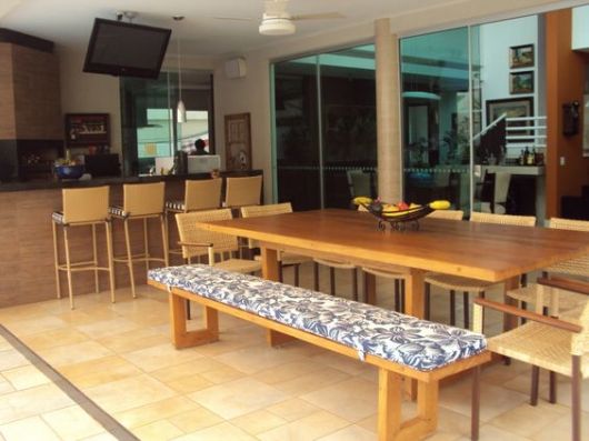 Foto de varanda gourmet espaçosa decorada com móveis de madeira como uma mesa de jantar, bancos e uma bancada ao fundo. 