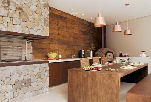 Foto de uma varanda gourmet com a churrasqueira feita de pedras e o resto do ambiente construído com madeira, como a mesa de jantar e a parede. 