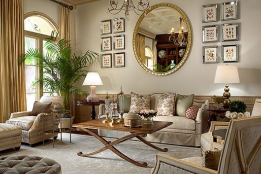 Sala com parede clean com quadros pequenos espelho redondo com moldura dourada, sofá grande com almofadas, mesa de centro em madeira.