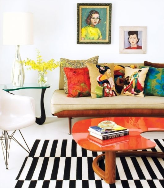 Sala clean na cor branca com quadros coloridos na parede, mesa de centro laranja, tapete listrado em preto e branco e sofá de três lugares com muitas almofadas.