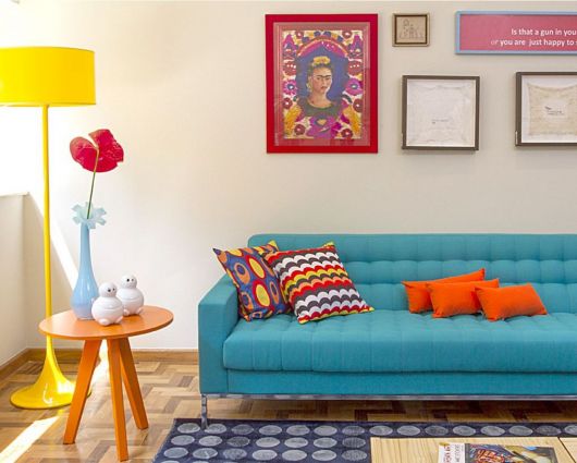Sala com paredes brancas, quadros coloridos, sofá azul e mesa lateral laranja.