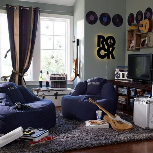 Sala de uma casa com dois puffs próximos a uma estante de televisão com discos de vinil, instrumentos musicais e um letreiro escrito "Rock" na parede. 