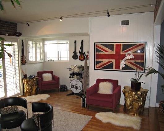 Sala de estar com duas poltronas, uma bateria do The Beatles, guitarras elétricas penduradas na parede e um quadro grande com a bandeira do Reino Unido. 
