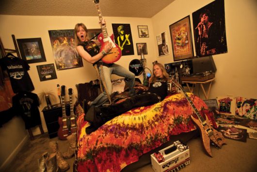 Casal em um quarto com decoração rock. Na parede há diversos posteres de bandas de rock famosas e diversos instrumentos estão posicionados no chão. 