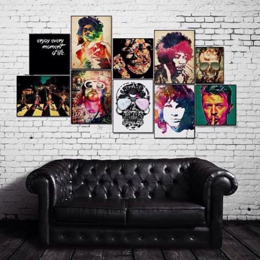 Sofá de uma sala de estar posicionado em frente a uma parede de tijolos repleta de quadros com ícones do rock como David Bowie, Jimi Hendrix e The Beatles. 