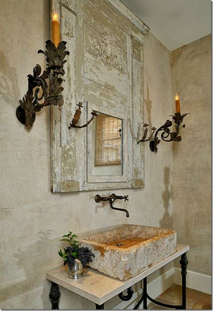 Foto da parede de um lavabo com duas velas, um espelho desgastado, uma cuba igualmente desgastada e uma torneira com formato sinuoso. 