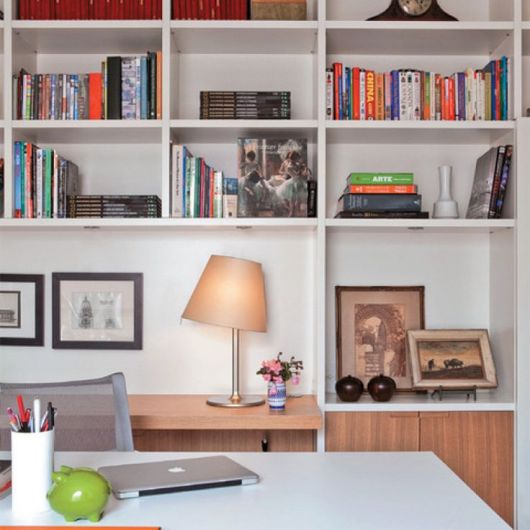 Foto de uma mesa com um notebook fechado sobre ela. Atrás da mesa há uma estante em MDF com quadros e livros. 