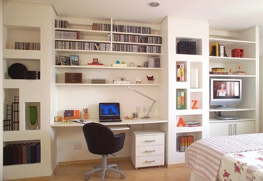 Foto de um quarto onde há uma parede totalmente voltada para Home Office.  Nela há uma cadeira em frente a um notebook e diversas estantes paralelas acima contendo objetos de escritório e decoração. 