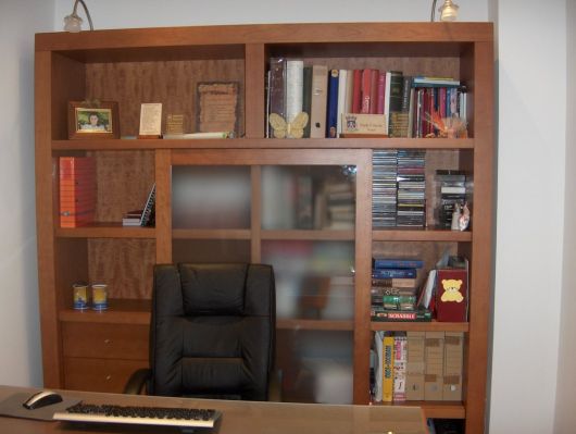 Foto de uma estante de madeira em frente a uma mesa e uma cadeira. Ela possui divisórias grossas e todas elas estão preenchidas com livros. 