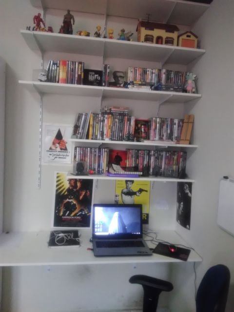 Mesa de MDF com um notebook e acima dela diversos prateleiras com DVDs e objetos de decoração. 