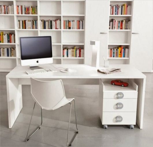 Mesa de escritório com um computador e uma cadeira em frente a ele. Na parede ao fundo há uma estante em MDF com diversos livros. 
