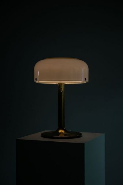 Foto de uma luminária que se parece com um abajur. Ela está em uma mesa em um ambiente escuro e não está acesa;.