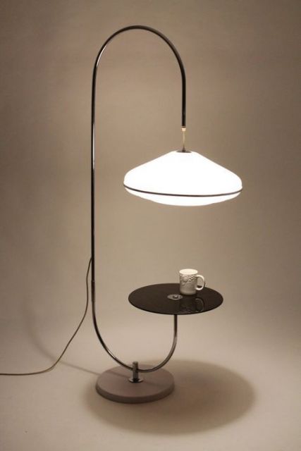 Foto de uma luminária que tem apenas uma linha sinuosa como base de sustentação. Em uma das pontas está uma plataforma reta que pode servir de apoio a objetos e na outra está uma lâmpada forte. 