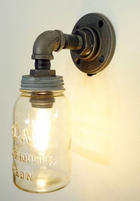Foto de uma luminária pendurada na parede. A sustentação é feita a partir de um material metálico e a lâmpada é coberta por um vidro. 