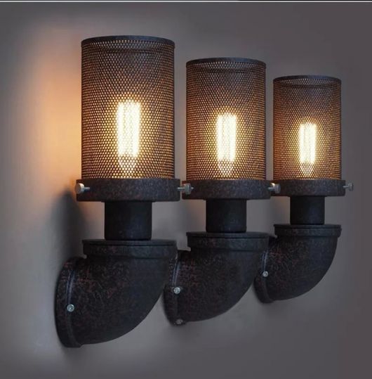 Foto de três luminárias iguais lado a lado. Elas estão voltadas para cima e seu base se assemelha a um cano. A lâmpada é coberta por uma estrutura de metal resistente que tem diversos furinhos. 