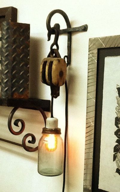 Foto de uma lâmpada coberta por um recipiente de vidro. Ela é ligada por meio de um fio a uma estrutura pendurada por um gancho em um objeto metálico pendurado na parede. 