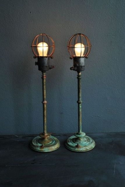 Foto de duas luminárias de chão penduradas lado a lado. Elas são altas e tem base de metal com visual retrô. As lâmpadas são grandes e potentes, protegidas por estruturas de metal. 