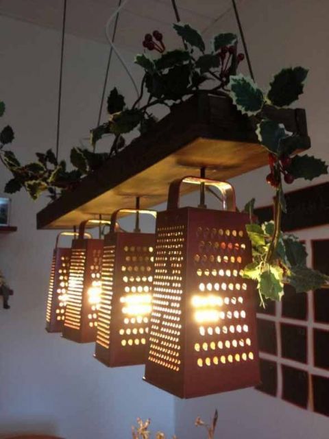 Foto de quatro lâmpadas enfileiradas penduradas em uma madeira suspensa no teto com pequenas folhas sobre ela. As lâmpadas estão dentro de raladores de queijo. 