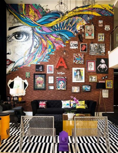 Foto de uma sala com parede de tijolos onde estão diversos quadros. Na parte alta da parede, ela se desfaz e dá lugar ao grafite do rosto de uma mulher com cabelo colorido. 