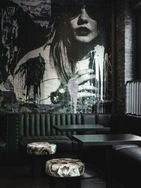 Foto de um ambiente com mesas de comer onde há um sofá e atrás dele o grafite com o desenho de uma mulher de óculos escuros