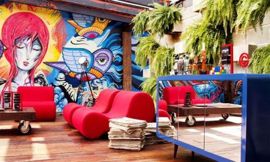Foto de um lounge com chão de madeira e puff. Em uma parede há o grafite de uma mulher e um pássaro, ambos com cores fortes e contrastantes entre si. 