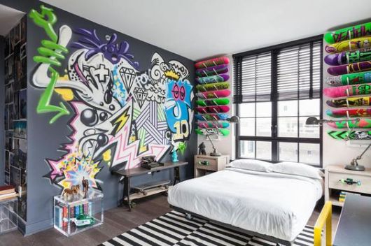 Foto de um quarto com a parede atrás da cama decorada com diversos shapes de skate. A parede ao lado da cama tem um grafite com diversos desenhos diferentes. 