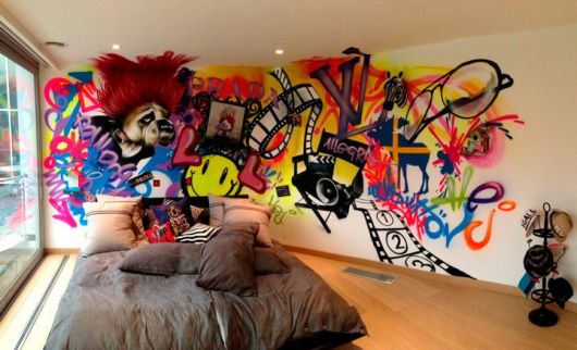Foto de um quarto com a parede atrás da cama decorada com grafite. Nos desenhos podemos observar rolos de filme, óculos, emojis e um bulldog. 