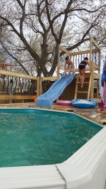 Crianças brincando em um escorregador para piscina de fibra que é fixado a uma estrutura de madeira onde elas sobre por meio de uma pequena escada. 