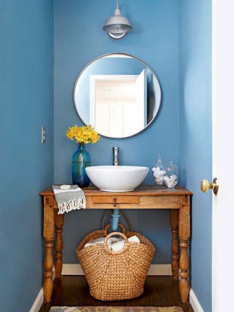 Banheiro com paredes azuis, espelho redondo e cesto embaixo da pia.
