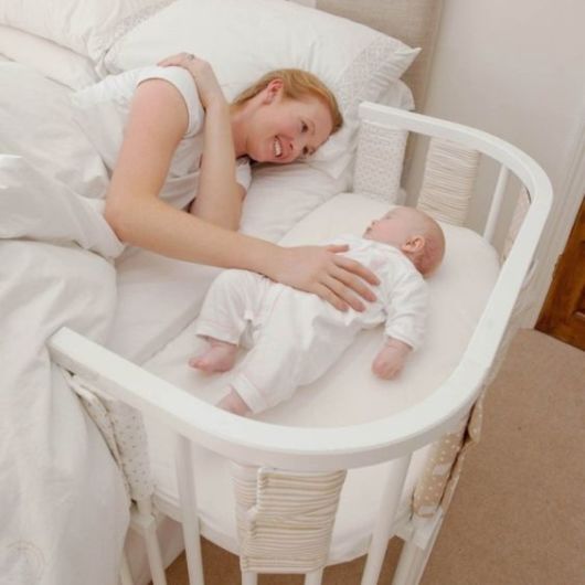 Mãe acariciando bebê que está em berço anexo à cama.